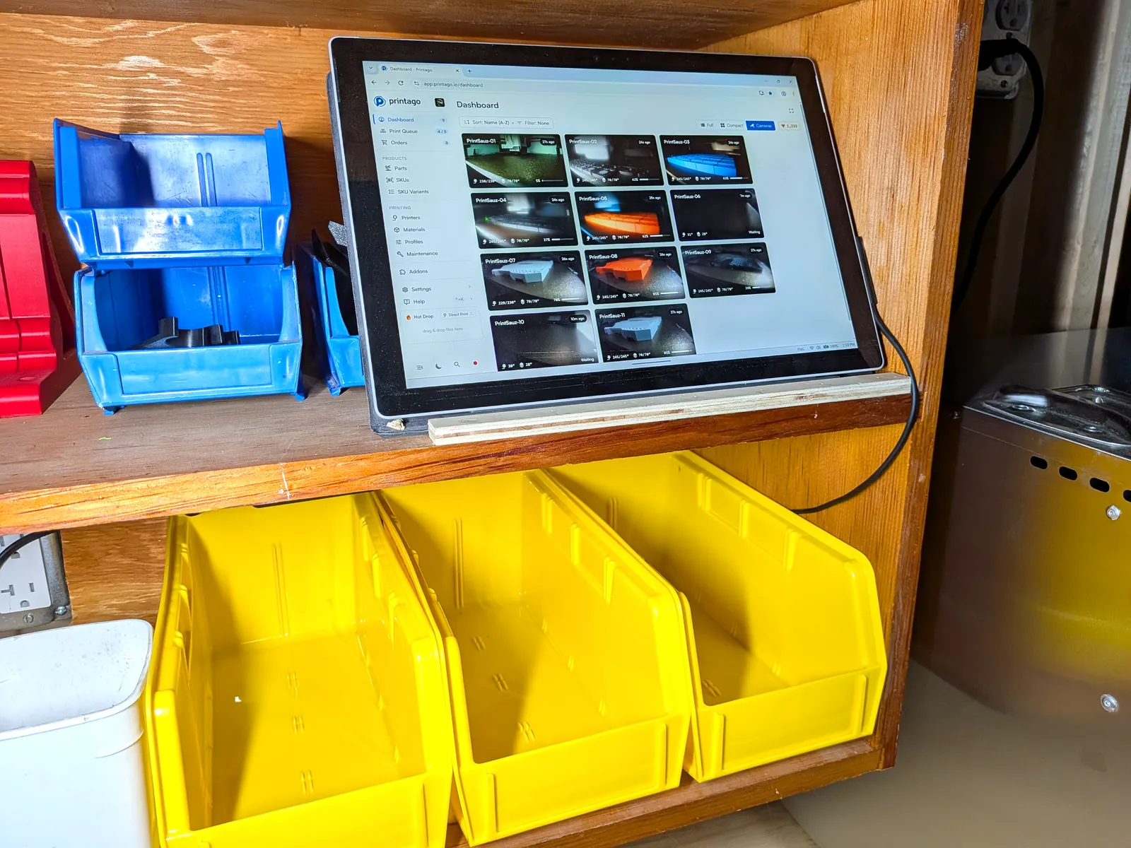 A tablet on the workbench showing the Printago dashboard with live printer camera tiles, next to parts bins.