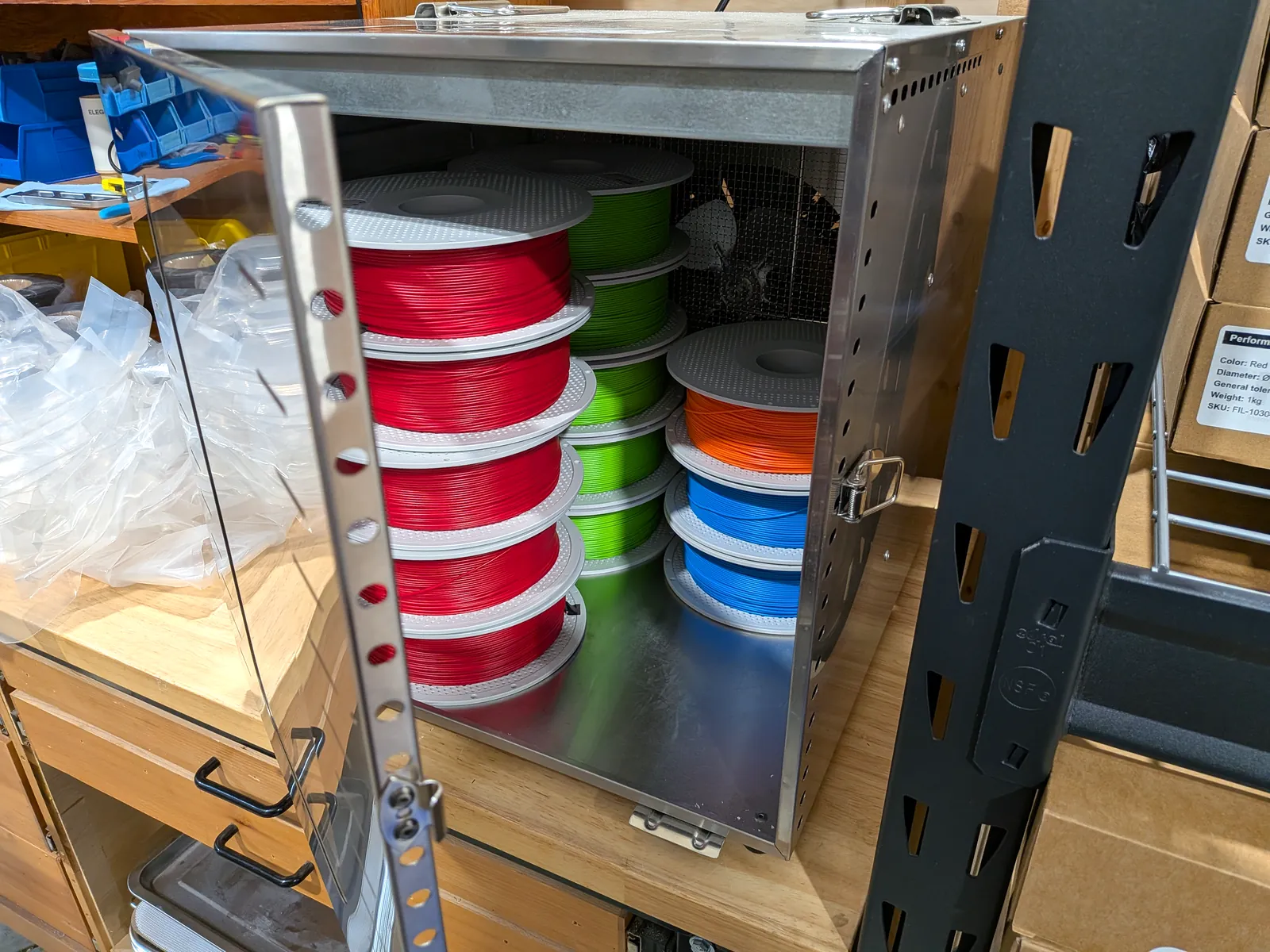 An open filament dry cabinet holding stacked spools of red, green, orange, and blue filament.