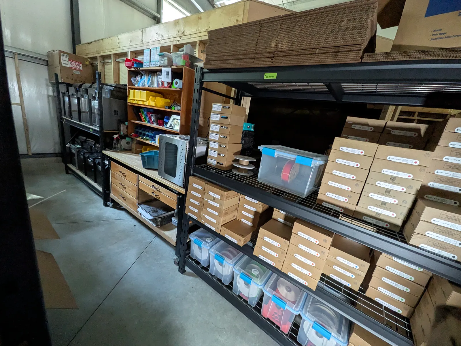 Warehouse shelving stacked with labeled filament boxes, spool stock, and packaging supplies.