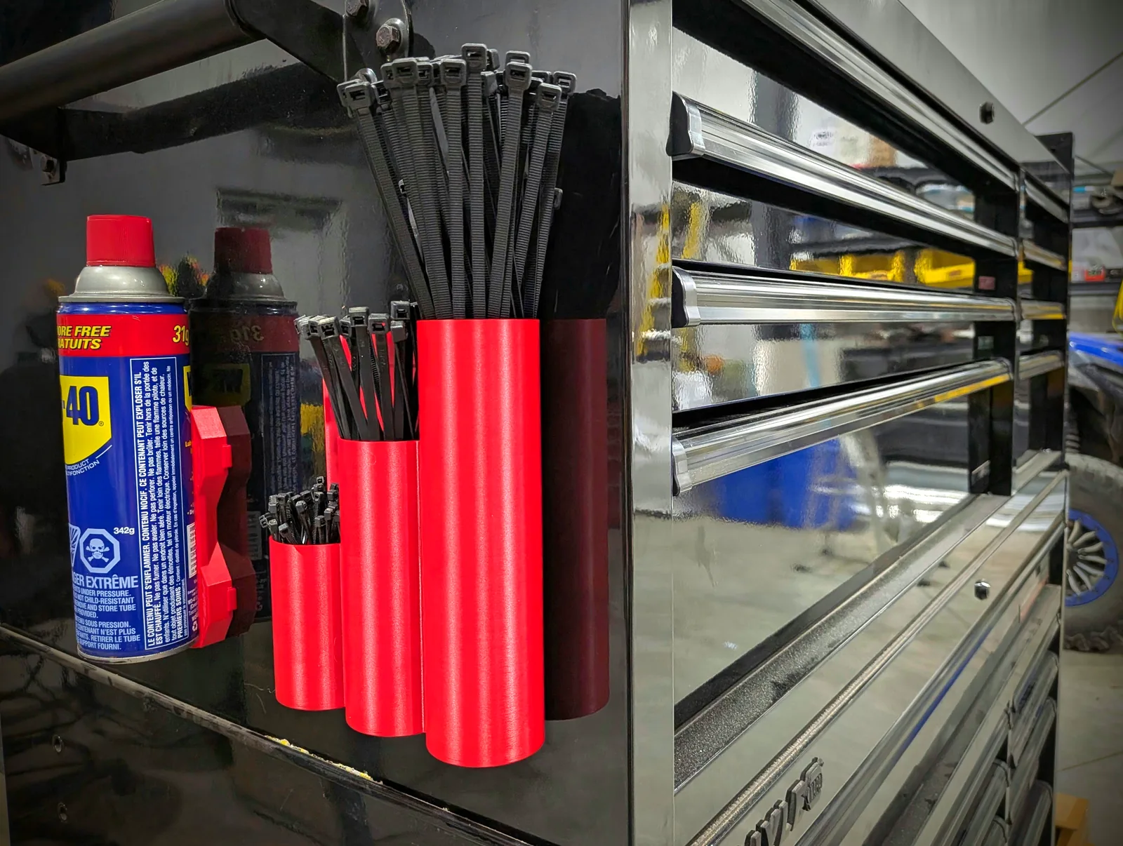 Red 3D-printed magnetic holders mounted on the side of a black tool chest, holding bundles of black zip ties next to spray cans.